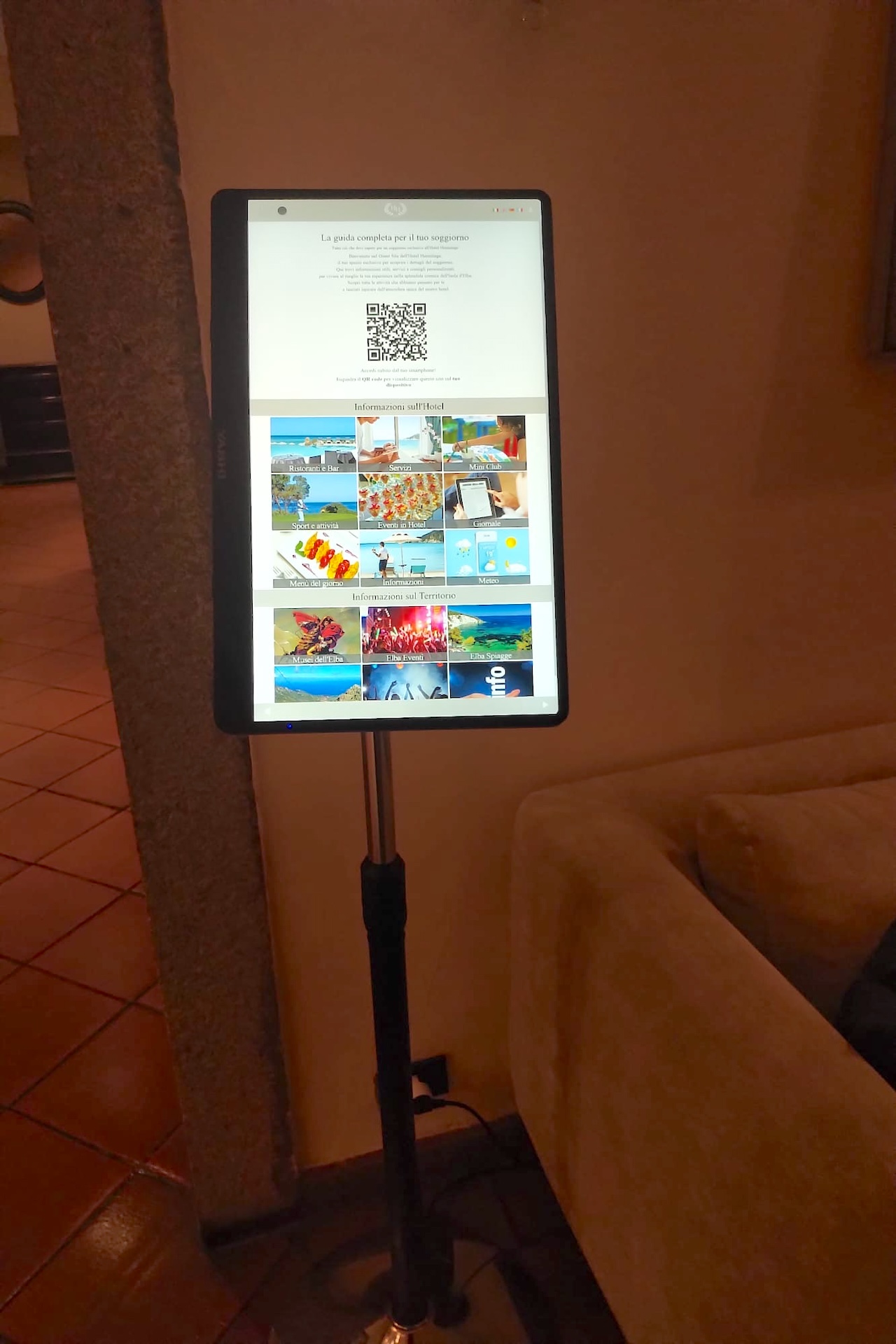 Totem informativo digitale con accesso al Guest Site per gli ospiti dell'hotel, posizionato accanto a un divano nella hall
